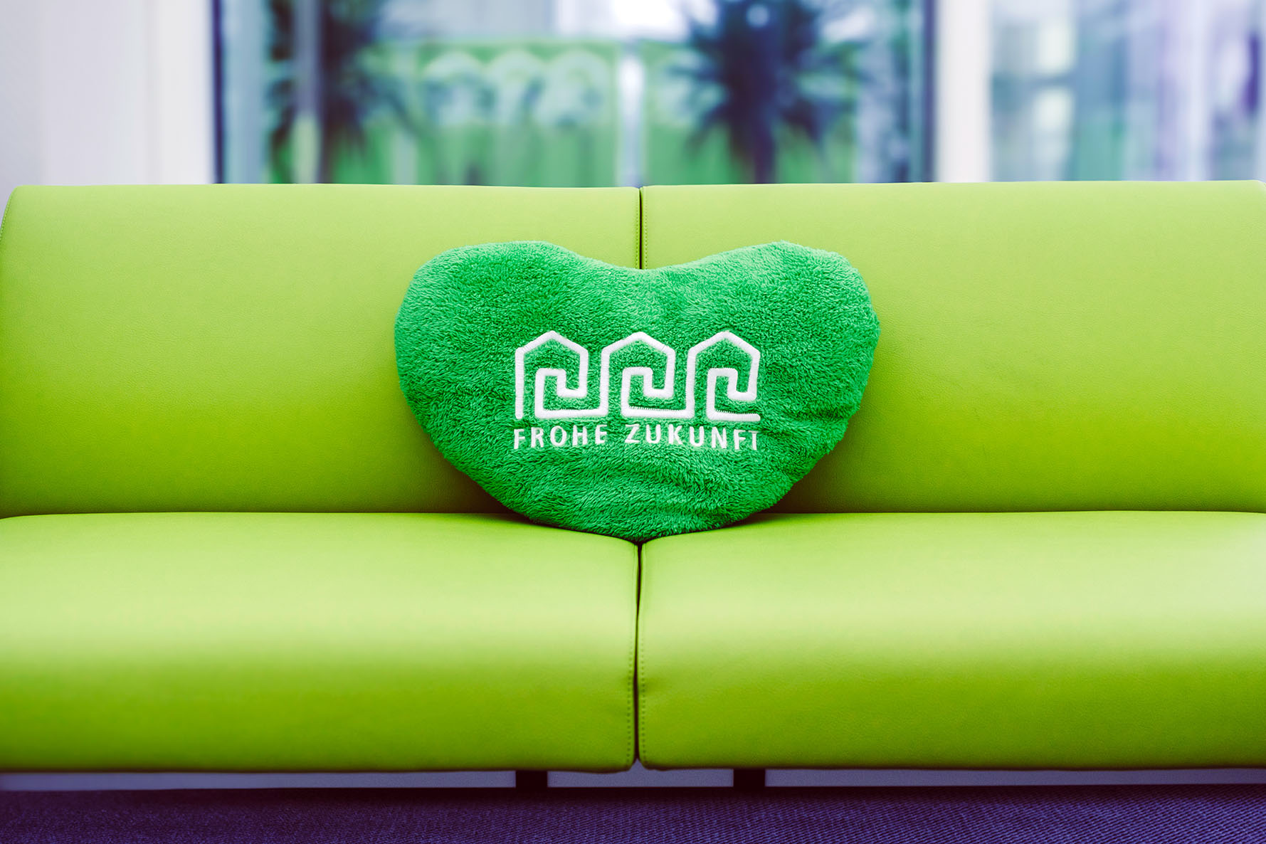 Grüne Couch mit grünem Stoffkissen in Herzform mit weißem Logo der FROHE ZUKUNFT Wohnungsgenossenschaft